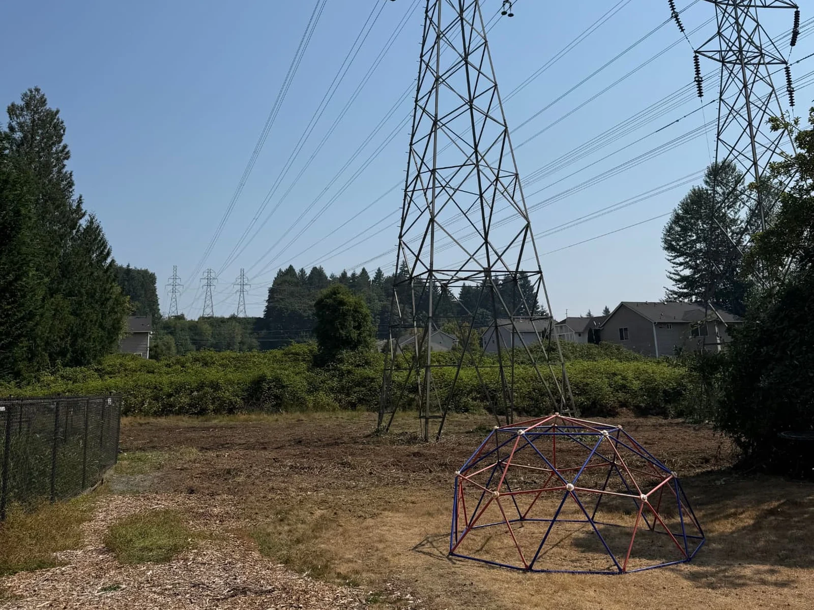 After: the same HOA common area cleared and brought to grade level, open bare ground ready for grass seed, with the fence, play dome and transmission towers unchanged